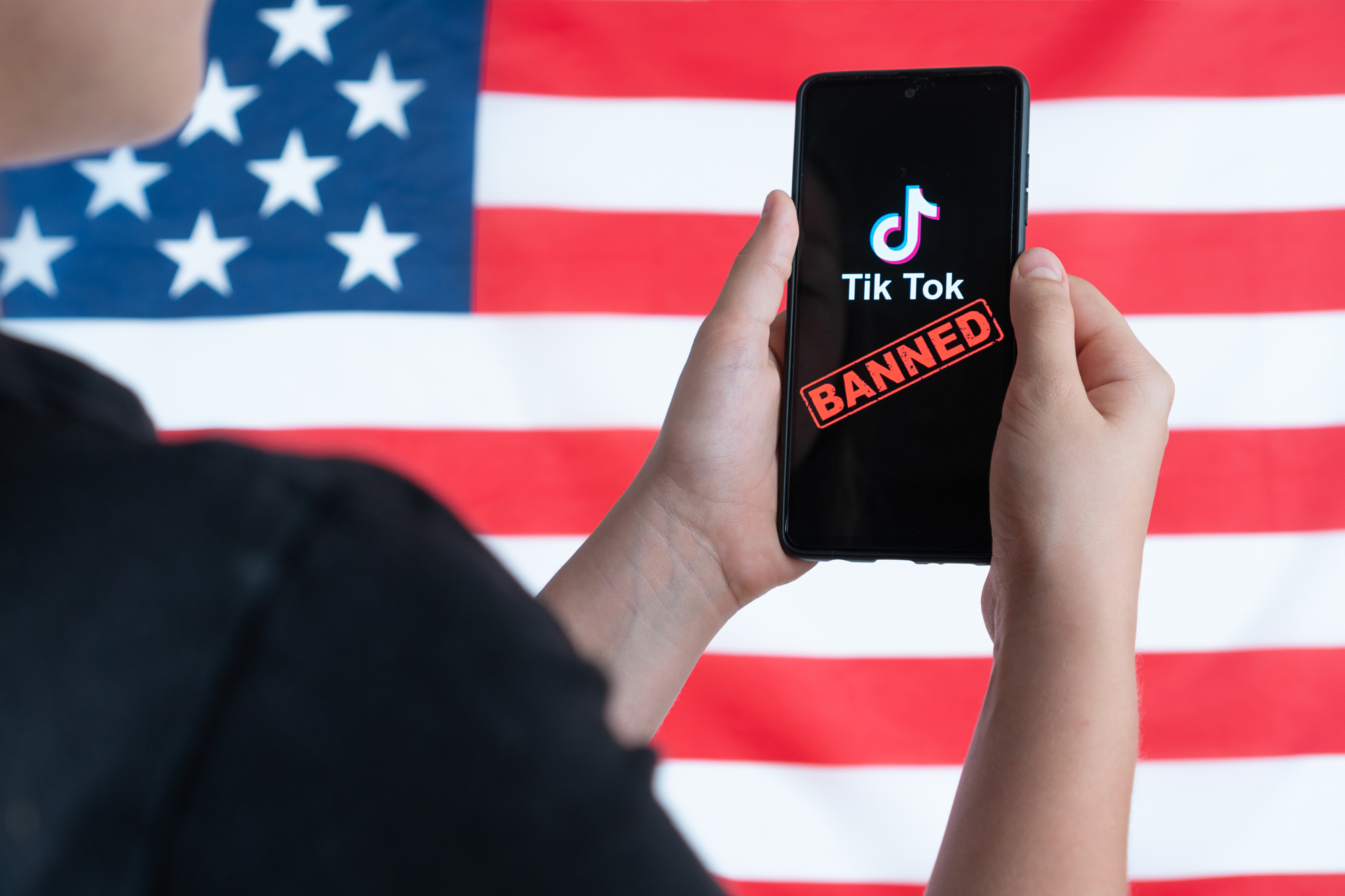 A teenager holds a smartphone with the Tiktok logo and against the background of the American flag. Selective focus. Donald Trump to ban Tiktok in United States. Russia, Kazan - August 08, 2020 tiktok